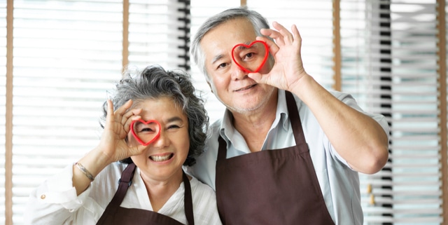 Asian senior couple wear brown aprons are holding red cookies cutters in heart shape over their eyes