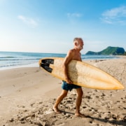 Older man holding a surfboard and walking on the beach after getting PRK treatment for his active lifestyle