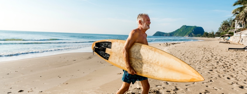Older man holding a surfboard and walking on the beach after getting PRK treatment for his active lifestyle